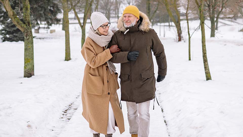 Senior couple walking arm in arm through a snowy park, dressed in warm winter clothing.