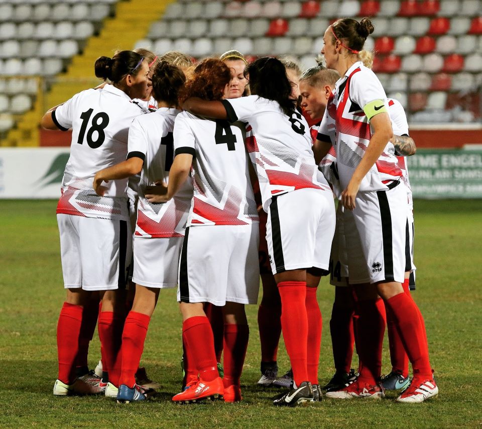 England Women’s Deaf Football Team huddling on the field in white uniforms with red accents and red socks.