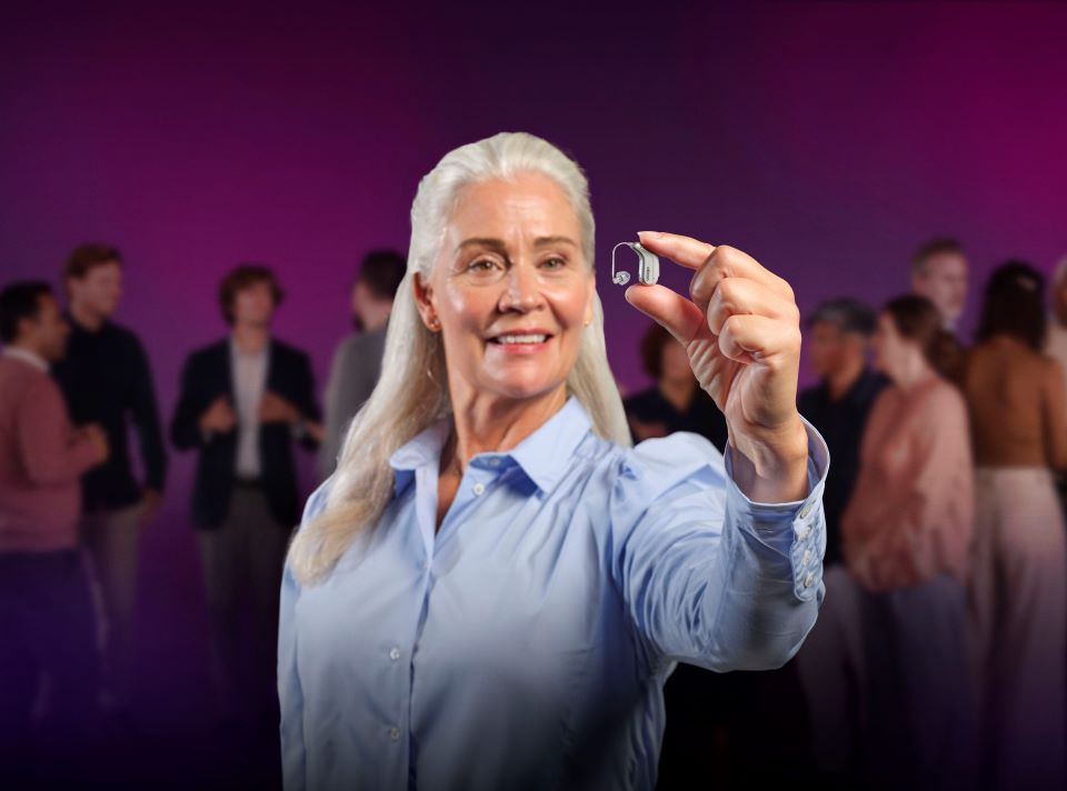 Senior woman holding a hearing aid in a social gathering setting.
