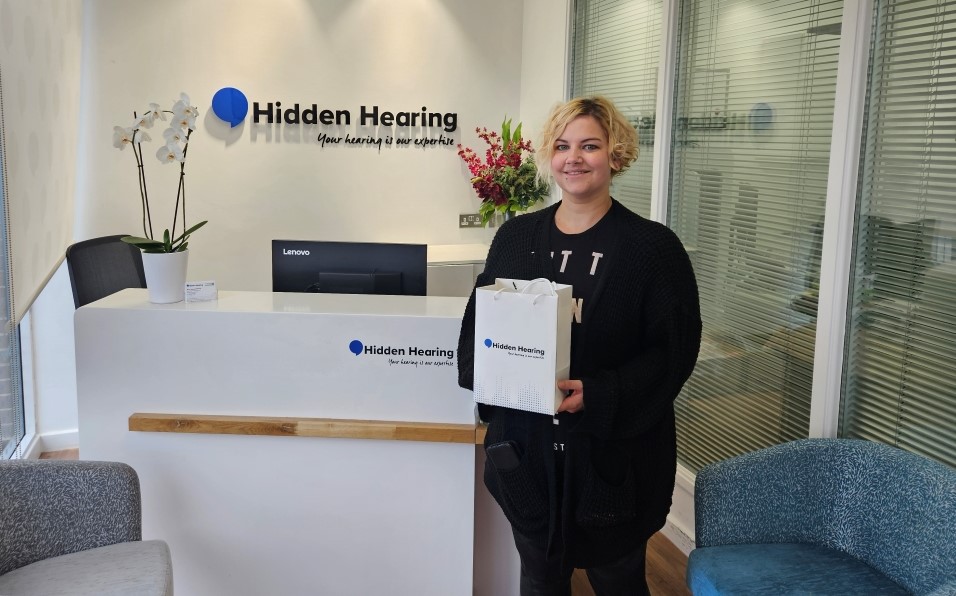 A woman standing at the reception area of Hidden Hearing clinic holding a branded bag containing her new hearing aids, with visible signage and welcoming decor in the background.