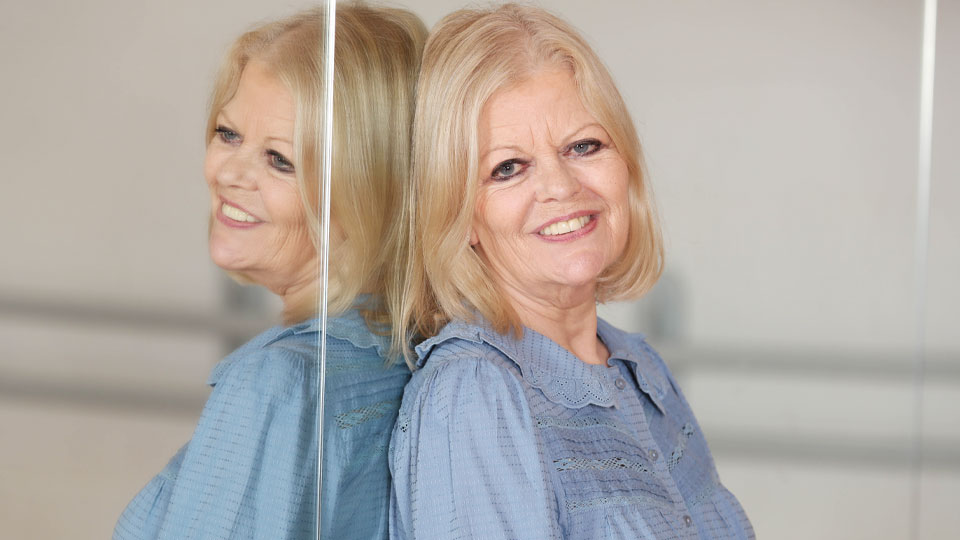 Elderly woman with blond hair wearing a blue blouse, leaning against a reflective surface.