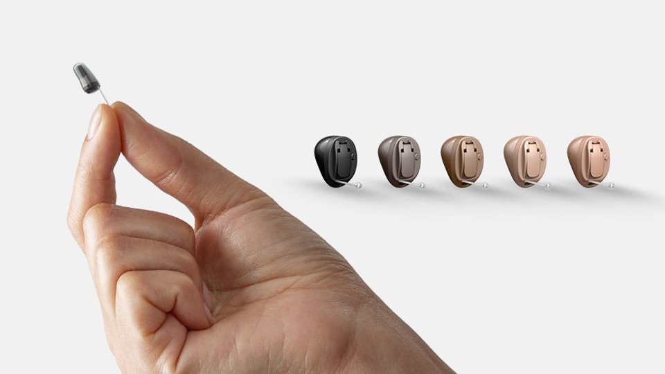 Oticon Own hearing aids displayed in multiple colors with a hand holding a small hearing aid tip for size reference.