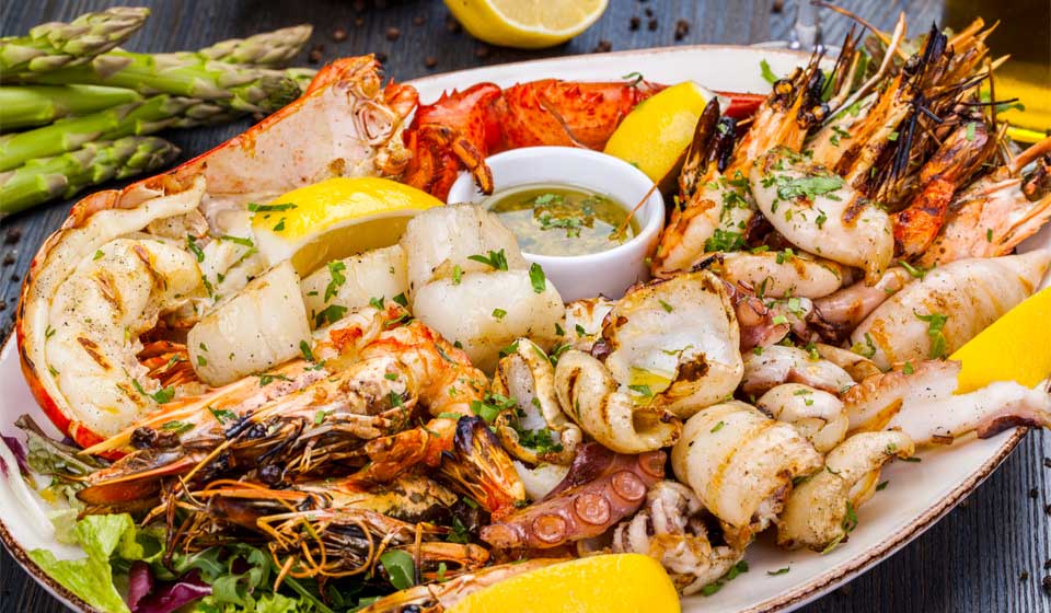 Grilled seafood platter with lobster, prawns, scallops, squid, and lemon wedges garnished with herbs, served with a dipping sauce and asparagus in the background.