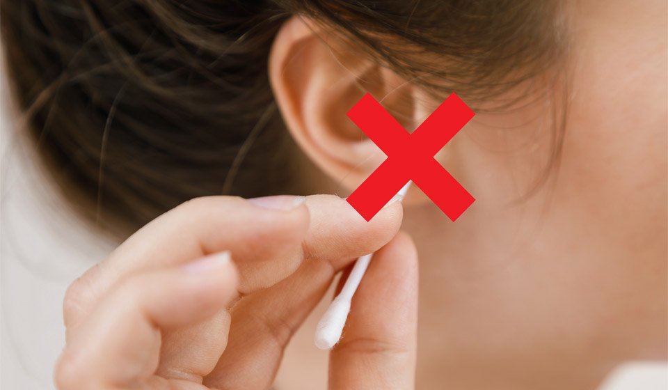 Girl inserting a cotton swab into her ear with a red cross symbol emphasizing improper ear cleaning.