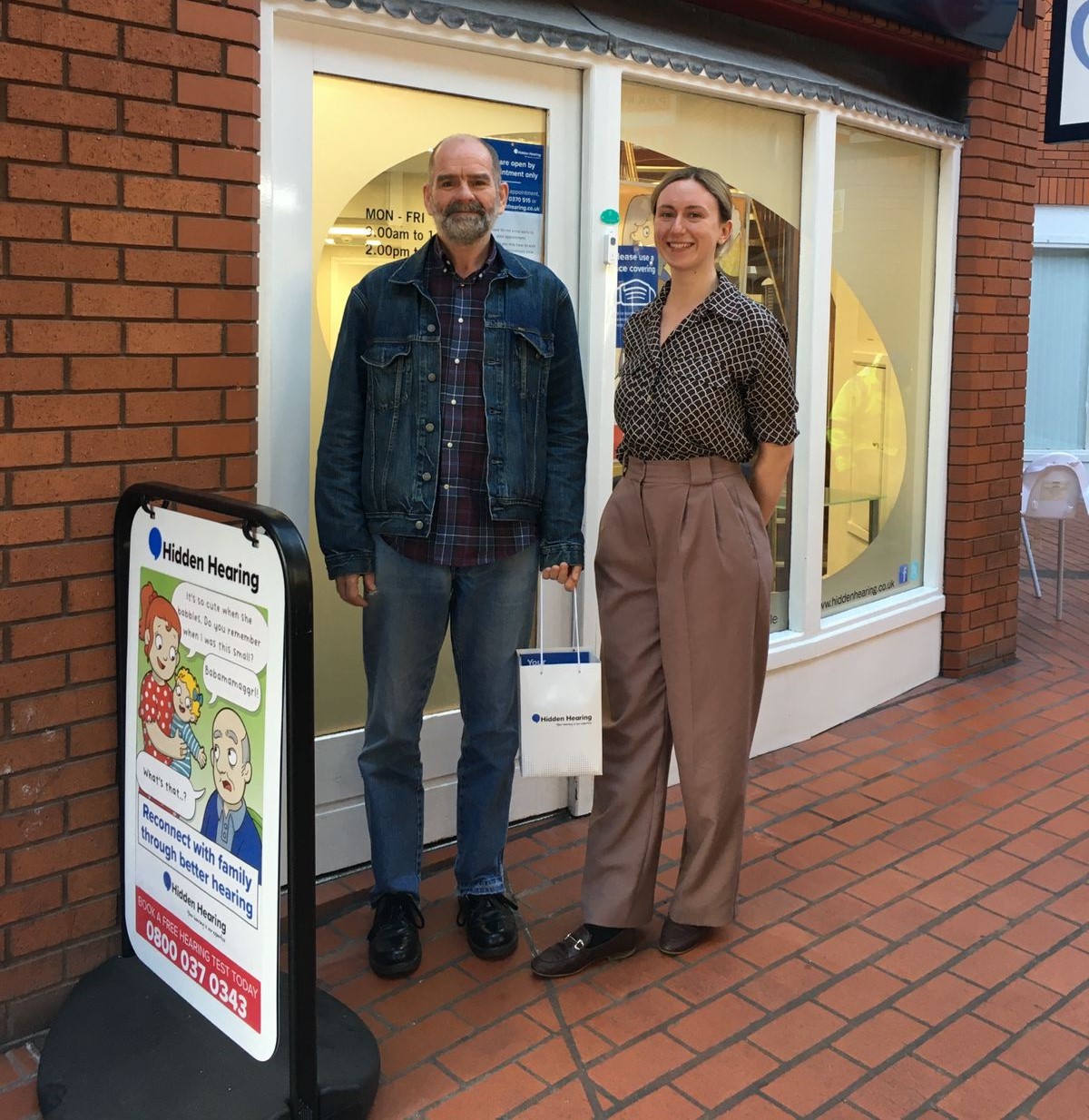 Customers outside a Hidden Hearing clinic holding a branded bag, with a promotional signboard nearby.