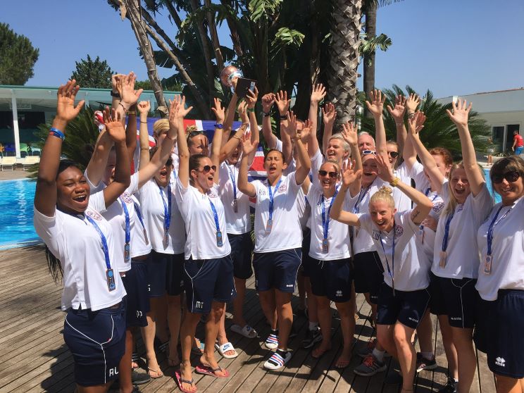 A group of individuals wearing matching white shirts and navy shorts raise their hands in celebration around a trophy. They stand on a wooden poolside deck with palm trees and clear skies.