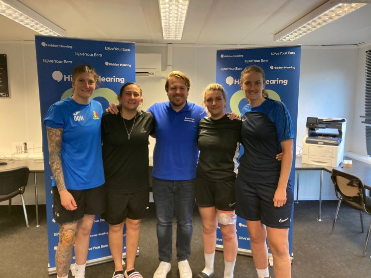 Five individuals stand together in a well-lit office environment between two 'Hidden Hearing' banners displaying the tagline 'Love Your Ears.' The space includes desks, chairs, and an office printer in the background.