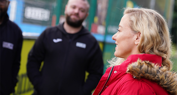 A woman in a red coat with fur detailing stands outdoors, interacting with two men in black athletic jackets. The background features a fenced area, blurred for depth and privacy.