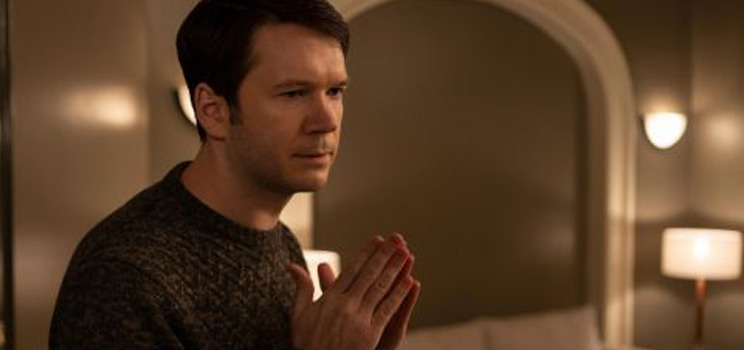 A person with a blurred face clasps their hands together in a warmly lit room featuring arched architecture, neutral walls, and softly glowing wall and table lamps.