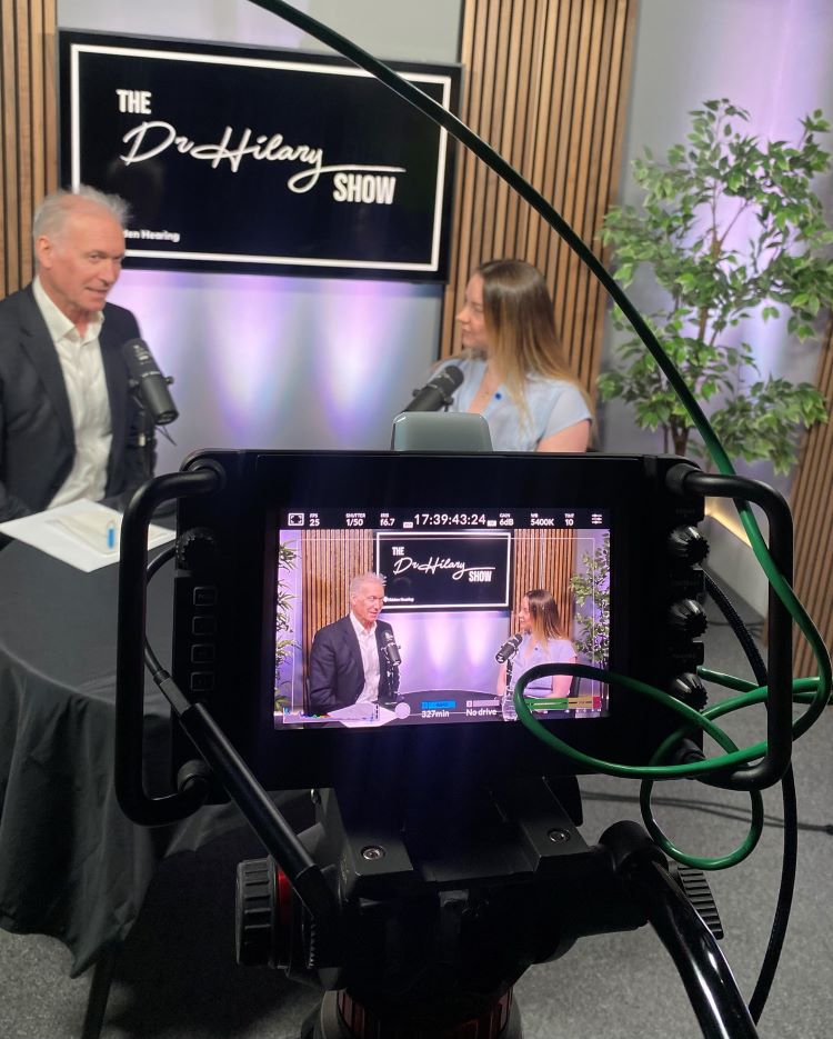 Dr Hilary hosting the Hidden Hearing Show with a guest in a professional recording studio setup.