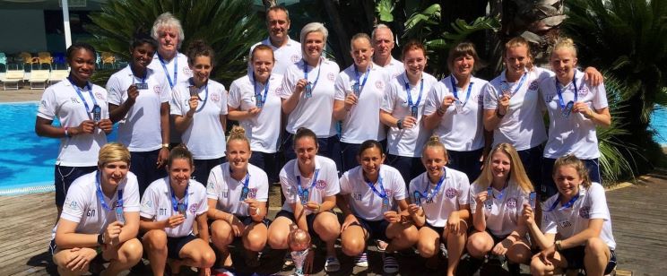 A team wearing white sports polo shirts with medals around their necks poses outdoors near a swimming pool and lush greenery, celebrating likely a sporting achievement.