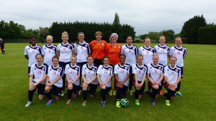 A soccer team of 18 players, wearing white and navy uniforms with visible numbers, poses outdoors on a grassy field. Two coaches in orange stand behind the kneeling team.