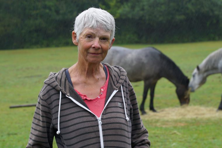 The description 'The impact of hearing loss and hearing aids at work' does not adequately describe this image. A more appropriate alt description might be: 'A person standing outdoors in a field with grazing horses in the background.'