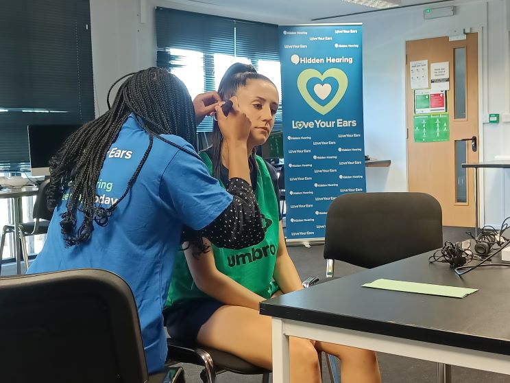 A hearing care specialist wearing a 'Love Your Ears' shirt examines a seated individual in a green Umbro bib, inside a hearing clinic featuring Hidden Hearing branding and promotional banners.