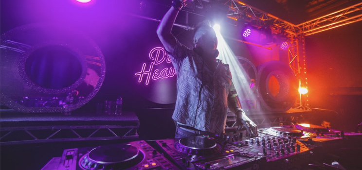 DJ performing under vibrant lights with a neon 'Deaf Heaven' sign in the background.