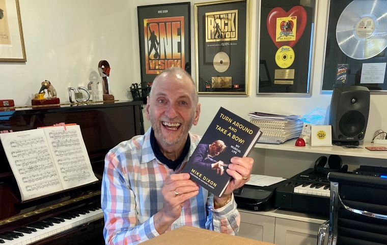 Renowned musical director celebrates improved hearing while holding an autobiography in a music-themed workspace.