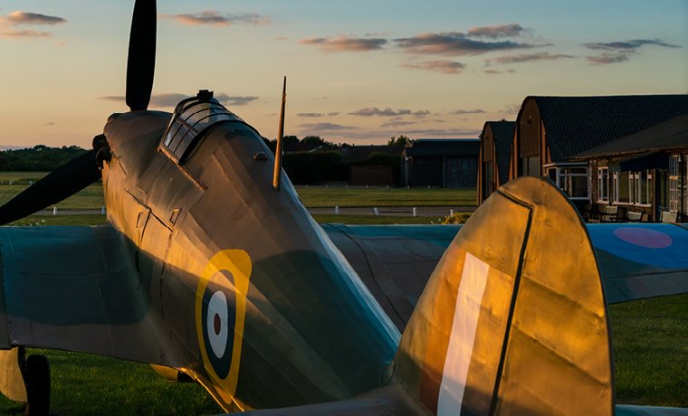 Spitfire plane parked on a grass airfield during sunset, with hangars and buildings in the background.