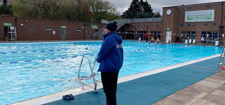 Tracey White standing by an outdoor swimming pool, wearing a blue jacket and observing swimmers.
