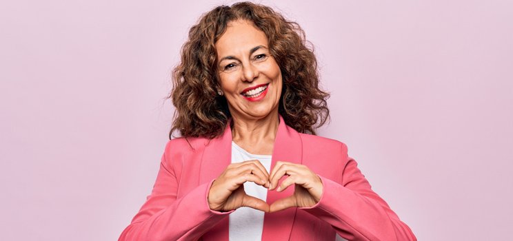 Woman in a pink blazer forming a heart shape with her hands against a light pink background.