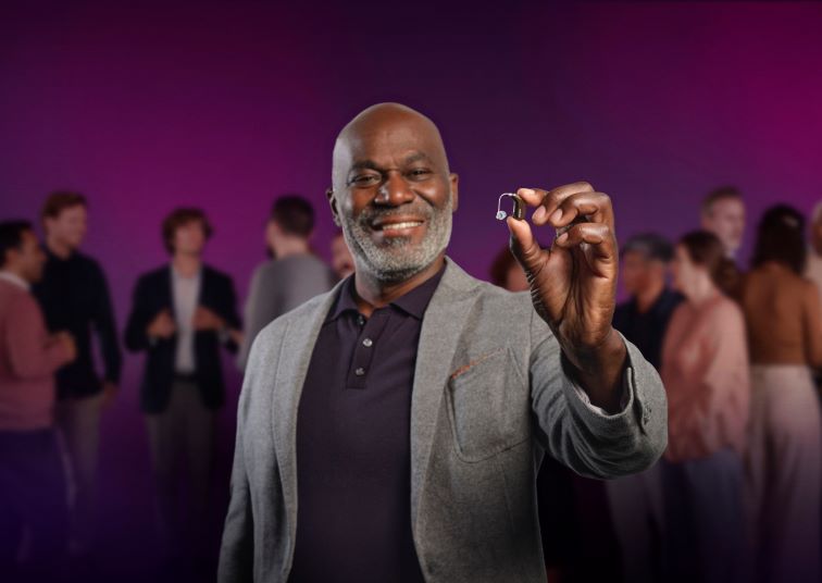 Oticon Intent hearing aid held in front of a group of people in a social setting.