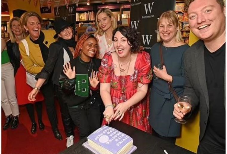 A group of people gathers at a Waterstones bookstore event; a person in a red floral dress cuts a purple frosted cake resembling a book, surrounded by shelves and promotional banners.