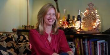 Woman in a red blouse sitting on a patterned couch in a festive living room decorated with lights, books, and holiday ornaments.