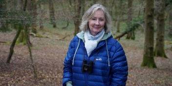 Woman in a forest wearing a blue jacket and binoculars around her neck.