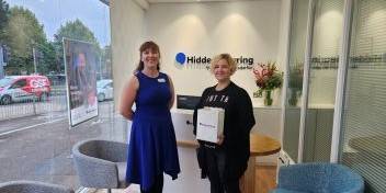 Woman smiling while holding a box containing new hearing aids, standing next to a hearing care specialist at a clinic.