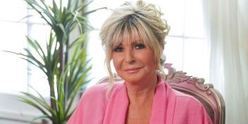 Senior woman wearing a pink robe, seated indoors with natural light and plants in the background.