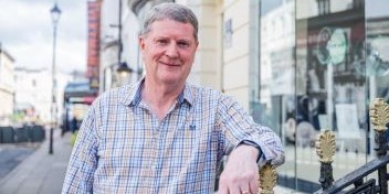 Senior man in a plaid shirt standing outdoors in a town center with shops in the background.