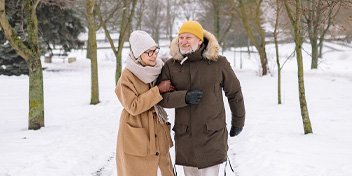 Elderly couple walking arm-in-arm through a snowy park, dressed in warm winter clothing.