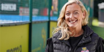 Blonde woman outdoors wearing a black jacket near a fenced area.