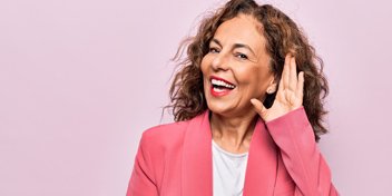 Woman in a pink blazer holding a hand to her ear, symbolizing listening and hearing awareness.
