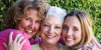Three women of different ages smiling and embracing closely outdoors, with greenery in the background.