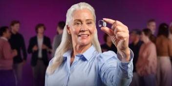 Senior woman holding a hearing aid with a blurred crowd in the background.