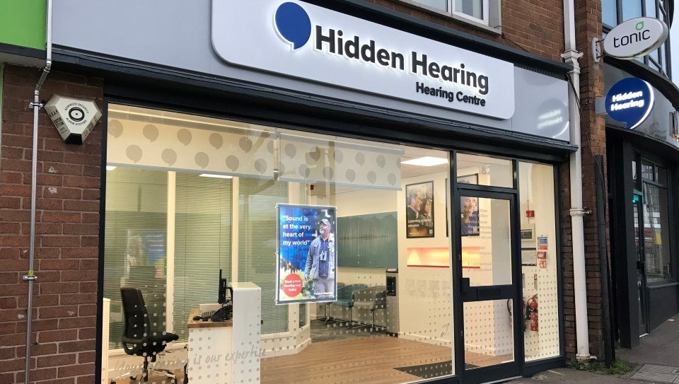 Hidden Hearing Exeter clinic exterior with visible signage, glass storefront, and interior view of the reception area.