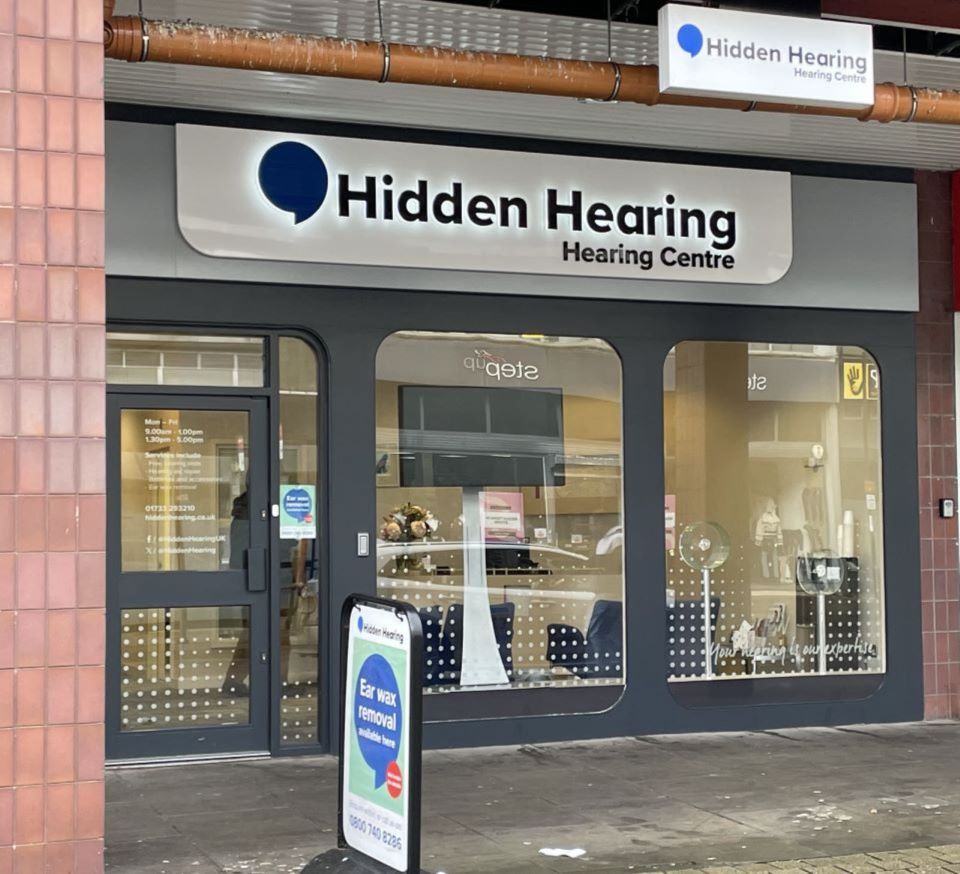 Hidden Hearing Peterborough clinic exterior featuring signage, large glass windows with dot patterns, promotional materials, and a sidewalk sign advertising ear wax removal services.