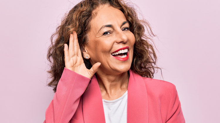 A person wearing a pink blazer holds a hand to their ear in a listening gesture, set against a solid light pink background, emphasizing hearing awareness or hearing care.