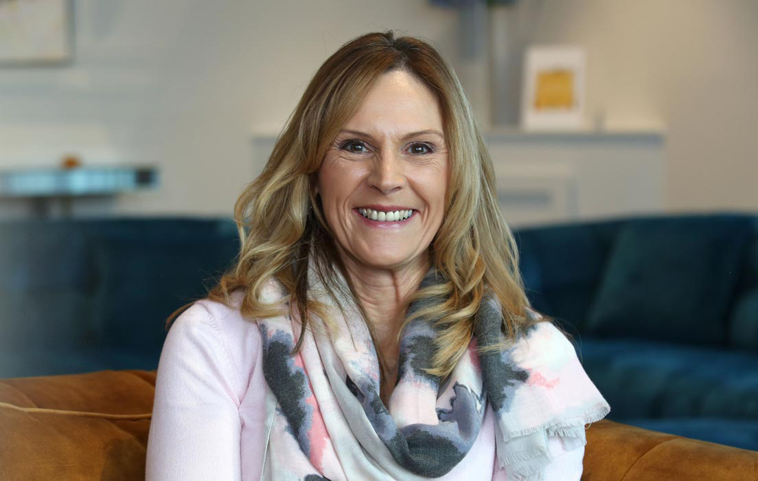 A woman with long blonde hair wears a pastel-patterned scarf and light pink sweater, seated on a brown sofa in a modern living room with a blurred blue couch and decorative background.