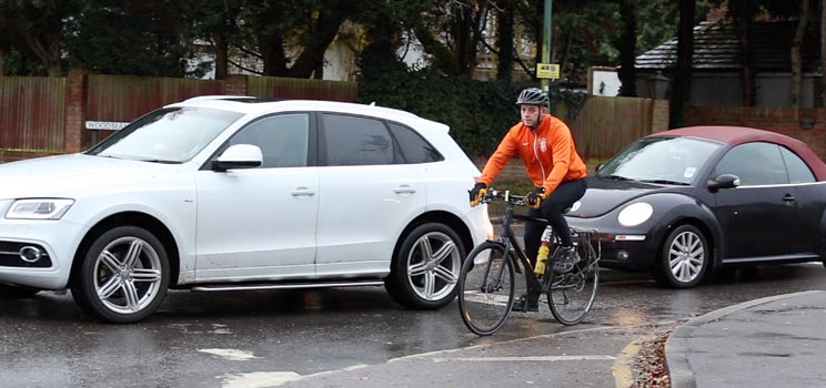 Hidden Hearing customer Alex Slight cycling on a road alongside parked cars.