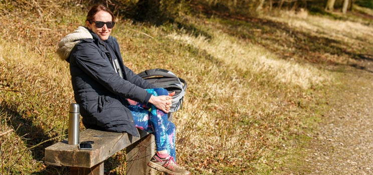 Helen Mol, a Hidden Hearing customer, sitting on a bench outdoors during a sunny day, holding a bag with a thermos and phone nearby.
