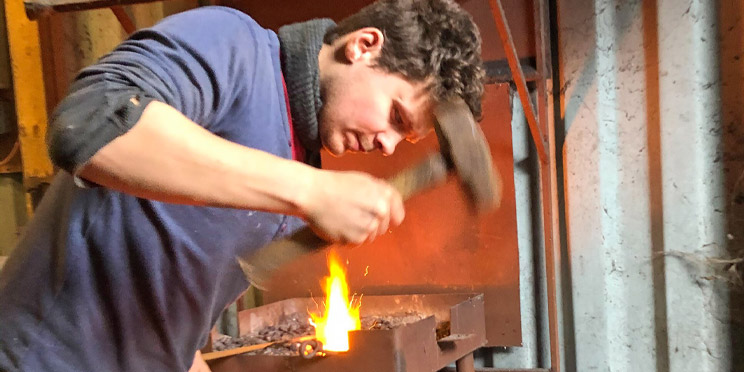Matthew Blackwell, a Hidden Hearing customer, forging metal with a hammer near a glowing forge.