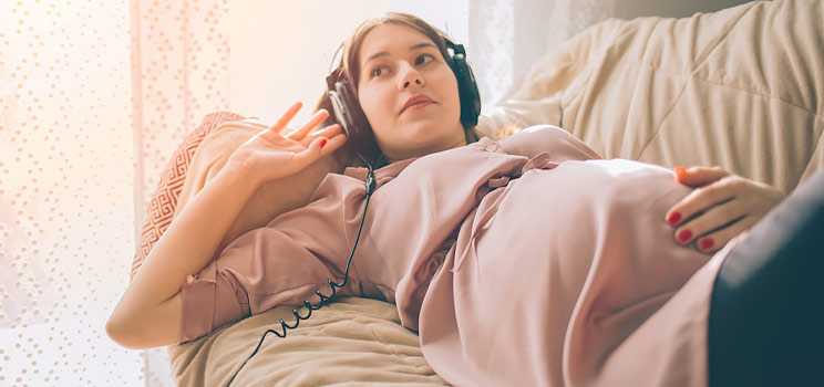 Pregnant woman relaxing on a sofa while wearing headphones and enjoying music.