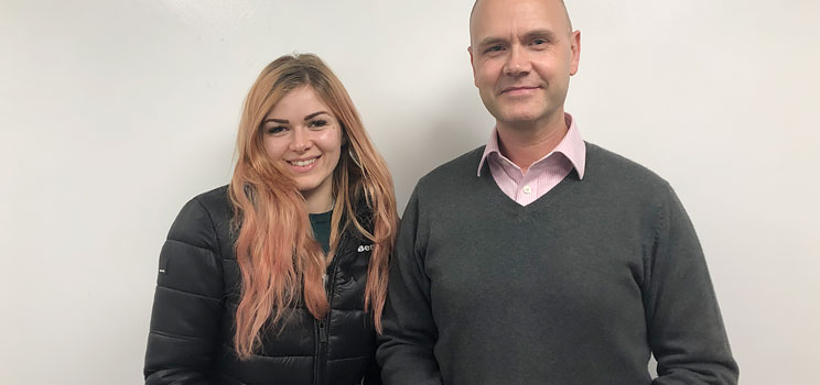 Hearing aid user Katie Lee stands next to Hidden Hearing Audiologist Darren Barnden against a white background.