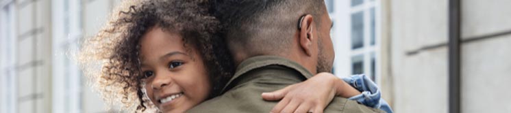 Man with short hair wearing a BTE hearing aid while carrying a child outdoors.