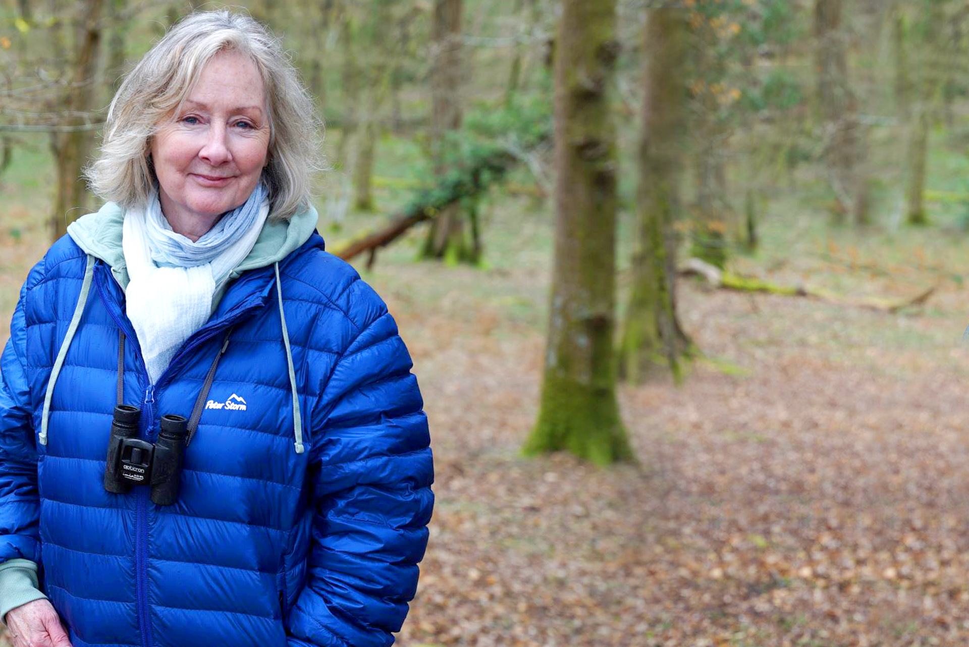 Woman in a blue jacket standing in a wooded area with binoculars around her neck.