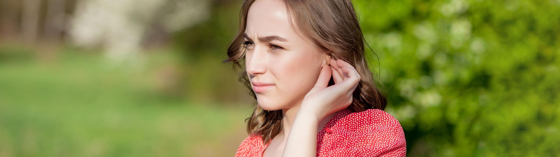 Woman adjusting or removing a hearing aid outdoors in a green, sunny setting.