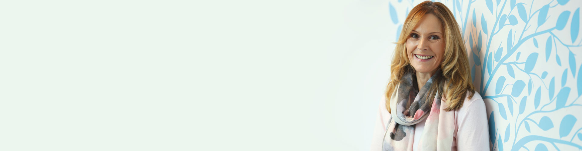 Sandra Pierce standing in front of a pastel blue patterned wall wearing a light scarf.