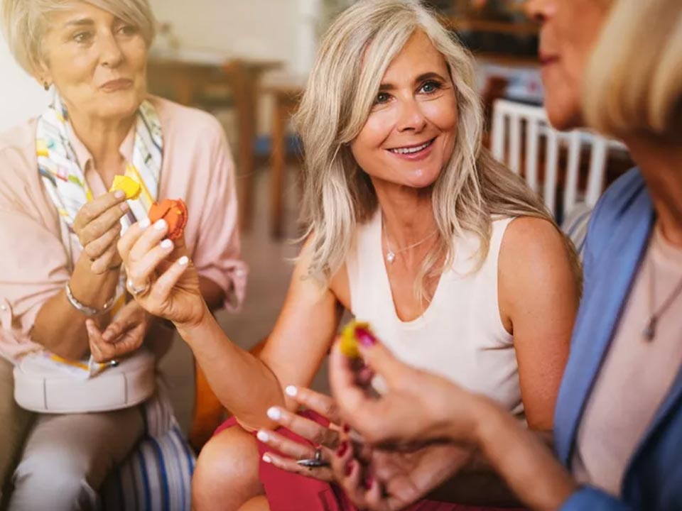 Three women are seated in a cozy, indoor setting, engaging in conversation while holding colorful macarons. The focus is on a woman with grey hair wearing a sleeveless top, gesturing mid-discussion. No visible hearing aids or text are present.