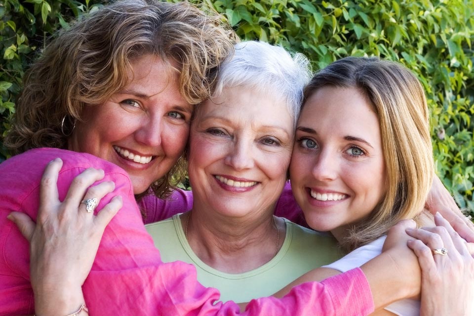 Three generations of women embracing outdoors in front of greenery.